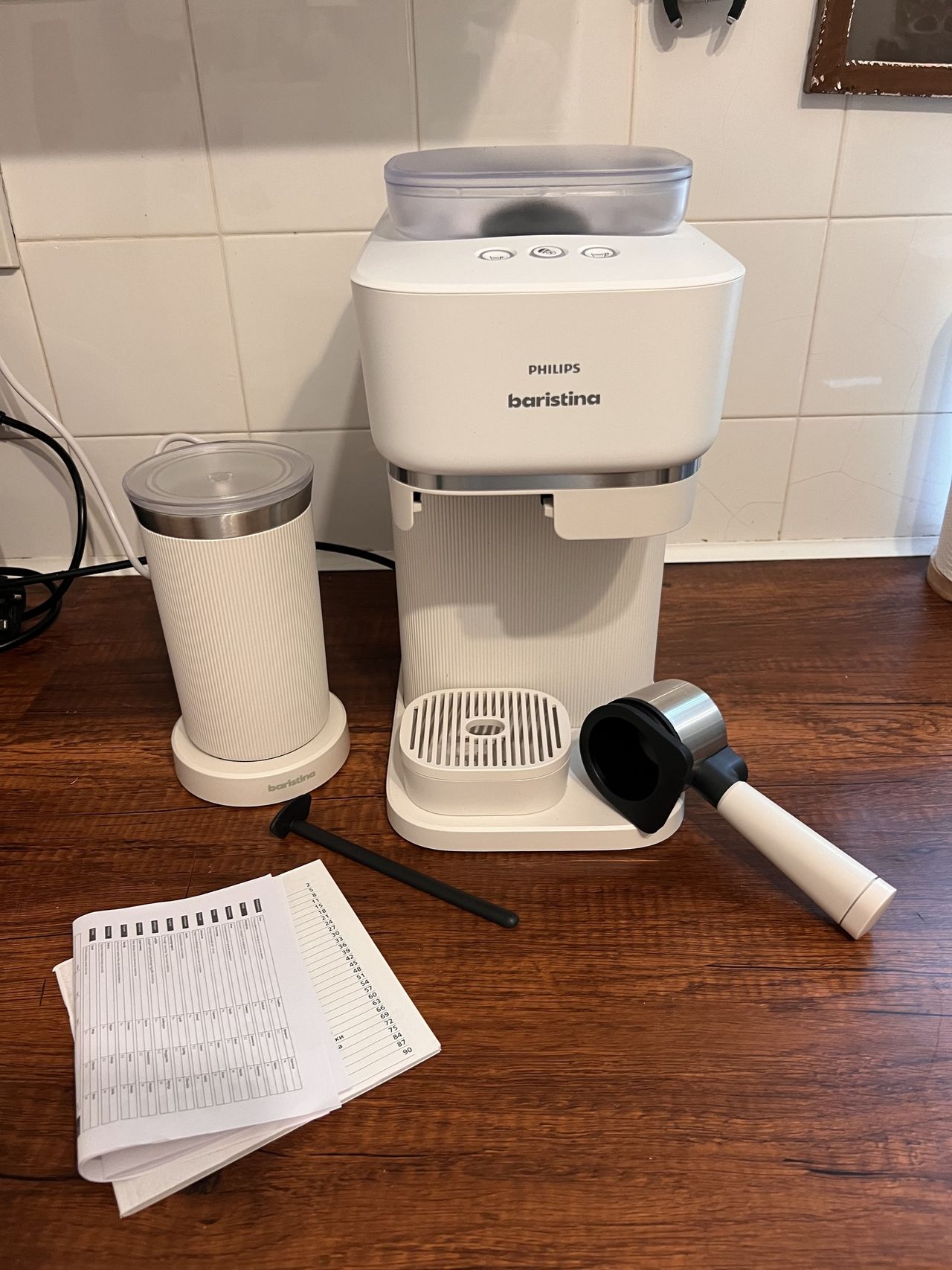 Image of a white Philips Baristina machine on a wooden countertop in a white kitchen with a milk frother, portafilter, and instruction manual beside it.