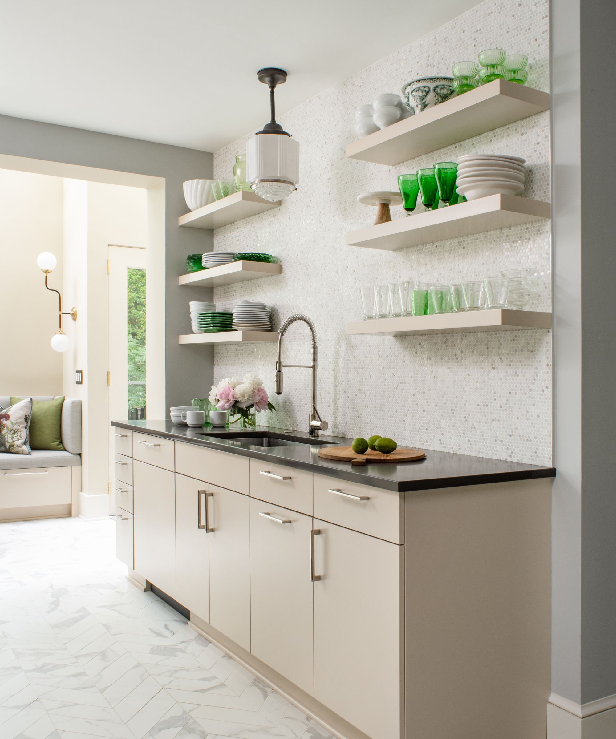 A neutral kitchen with white walls, wood cabinets, black countertops, and white floating wall shelves decorated with green glass decor and tableware