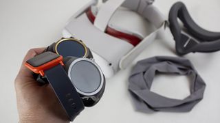 Several smartwatches held in a hand, with the Meta Quest 2 and some fitness equipment in the background.