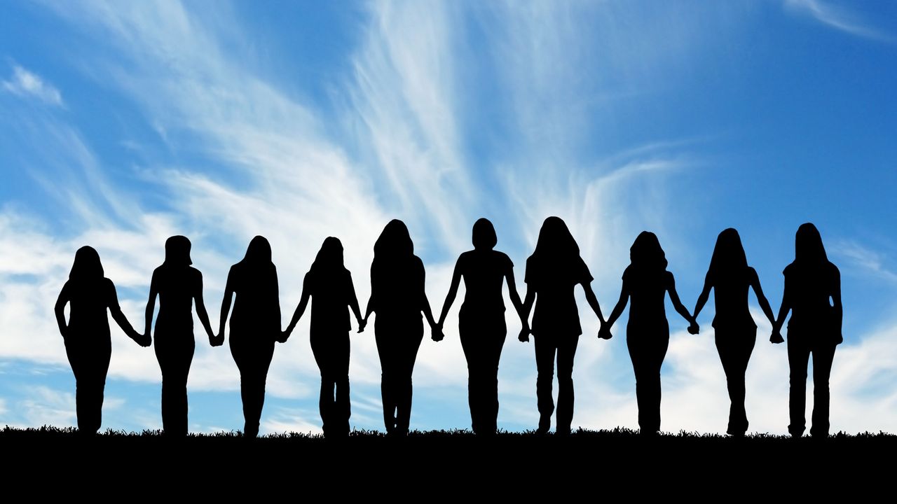 A line of 10 women holding hands against the backdrop of a blue sky, their backs to the camera.