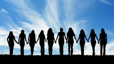 A line of 10 women holding hands against the backdrop of a blue sky, their backs to the camera.