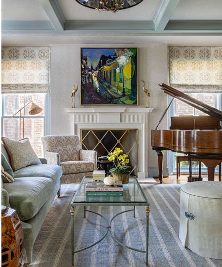 Traditional living room with grasscloth walls, large fireplace with gold grate and abstract landscape above with two gold birds on the mantel, a pale blue sofa, a glass coffee table, and a large piano