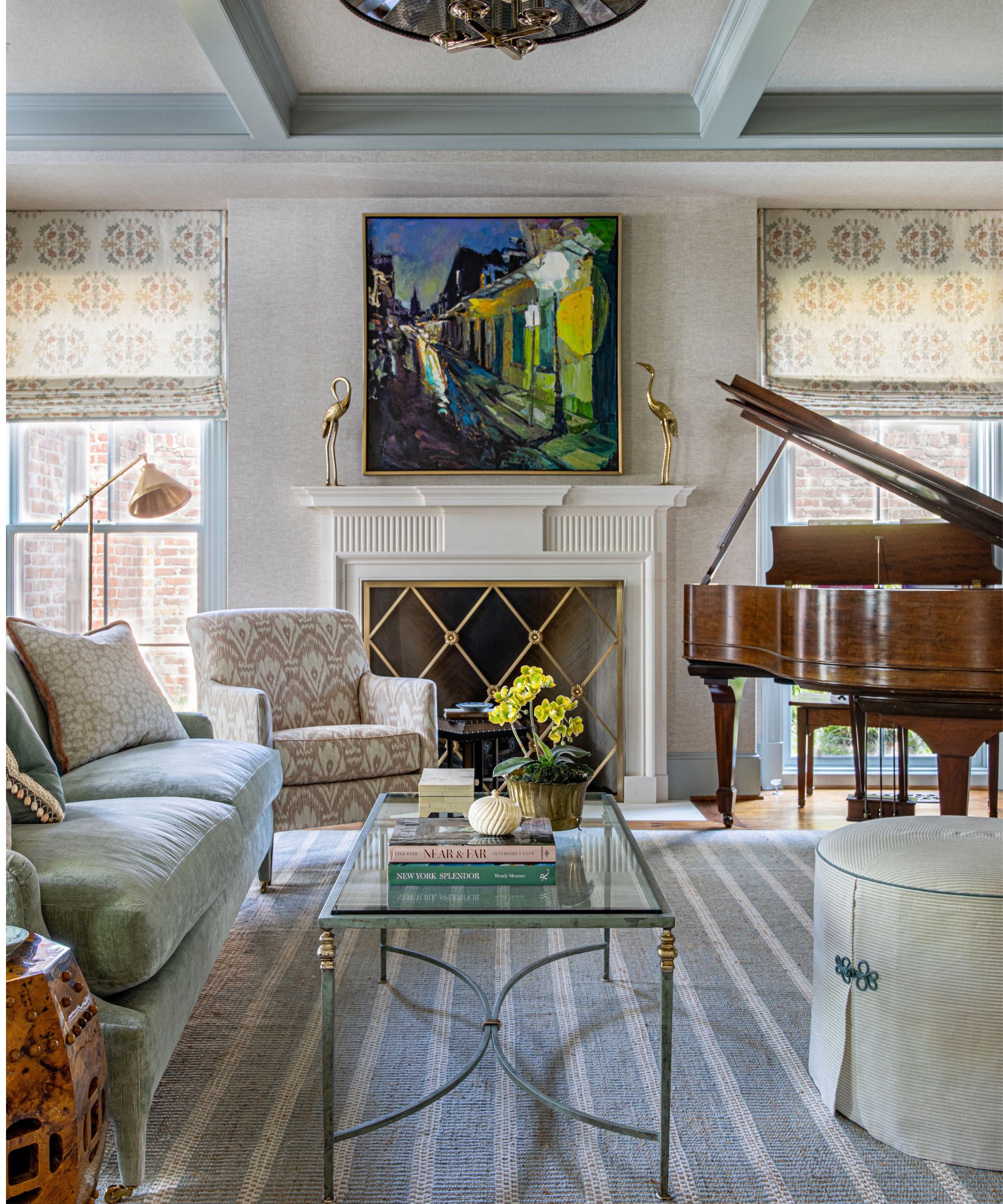 Traditional living room with grasscloth walls, large fireplace with gold grate and abstract landscape above with two gold birds on the mantel, a pale blue sofa, a glass coffee table, and a large piano