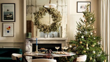 A dining room with cream walls, a large mirror with a wreath hanging in front, a marble fireplace, and a large Christmas tree in the corner. A round table and boucle dining chairs sit in the centre.