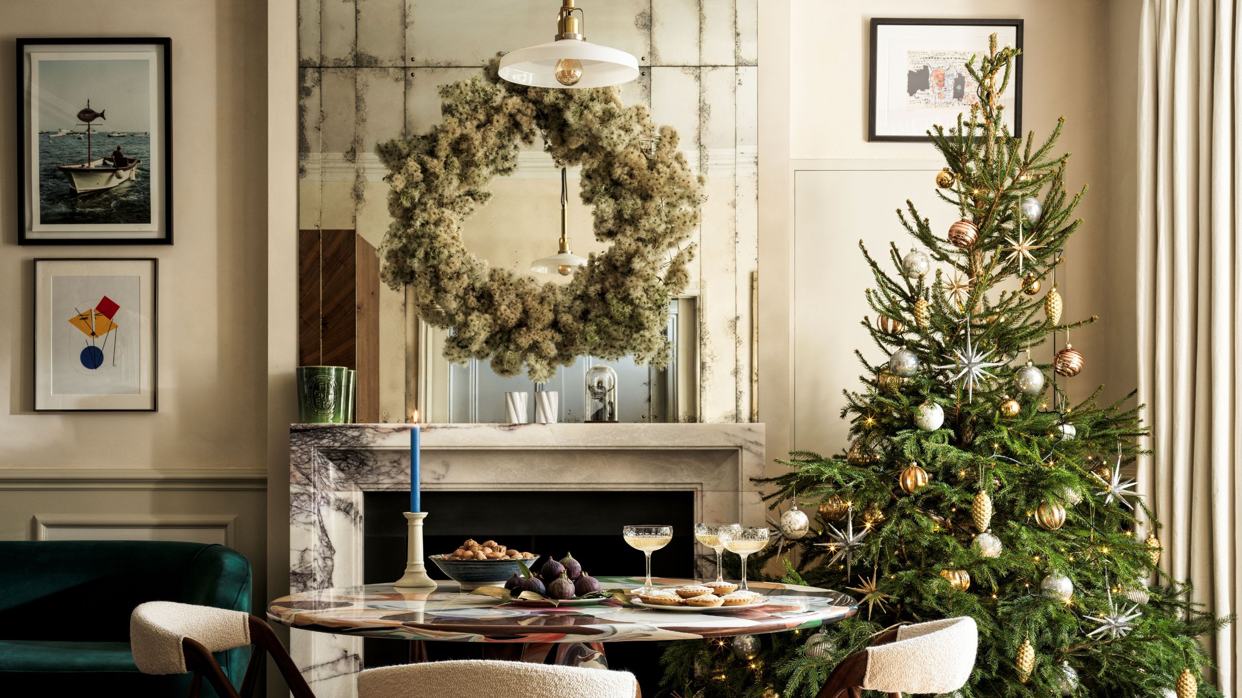 A dining room with cream walls, a large mirror with a wreath hanging in front, a marble fireplace, and a large Christmas tree in the corner. A round table and boucle dining chairs sit in the centre.