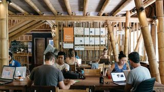 People work at laptops on wooden tables surrounded by bamboo pillars and under a high bamboo ceiling