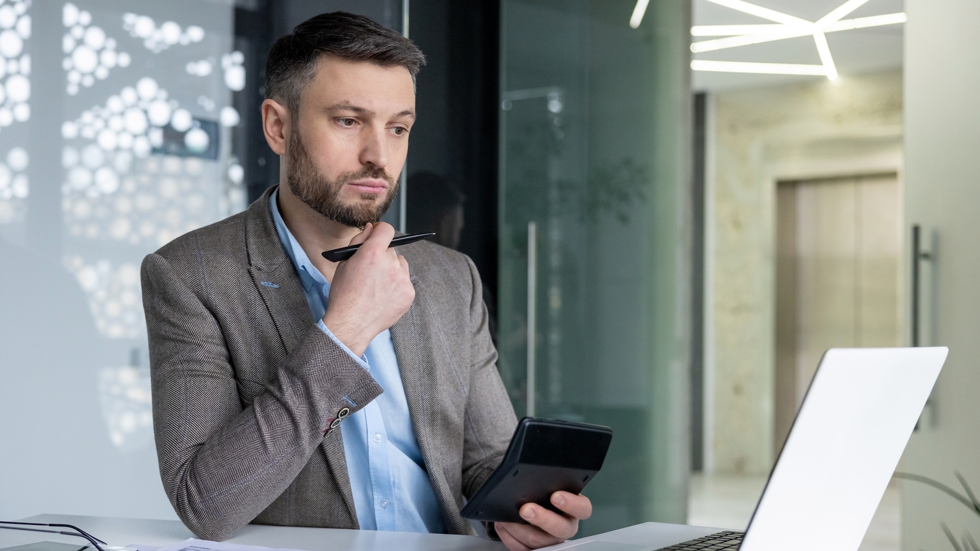 A thoughtful businessman holds a calculator while looking at his laptop.