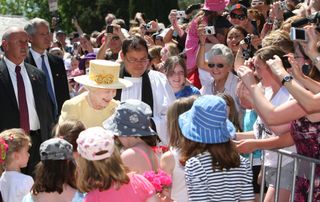 Queen Elizabeth in a yellow hat and coat greeting a huge crowd of fans taking photos of her