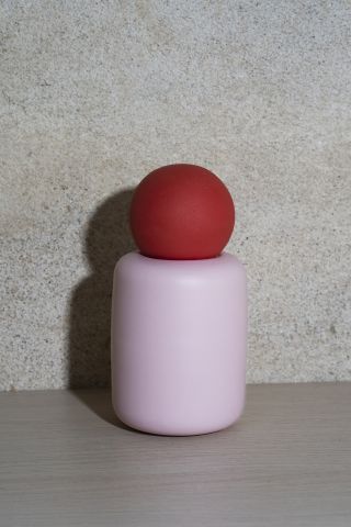 pink vase with red sphere on top placed on a wooden desk and photographed against a concrete wall