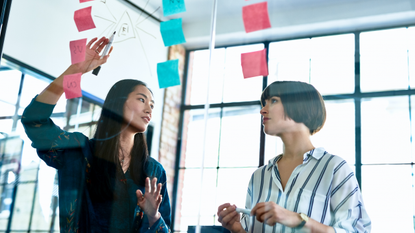 Two women discuss work in a conference room, using note cards and a clear glass board. 