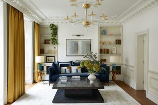 white living room with turmeric drapes, navy couch, large coffee table, bookcases, artwork, chandelier, rug, wooden floor