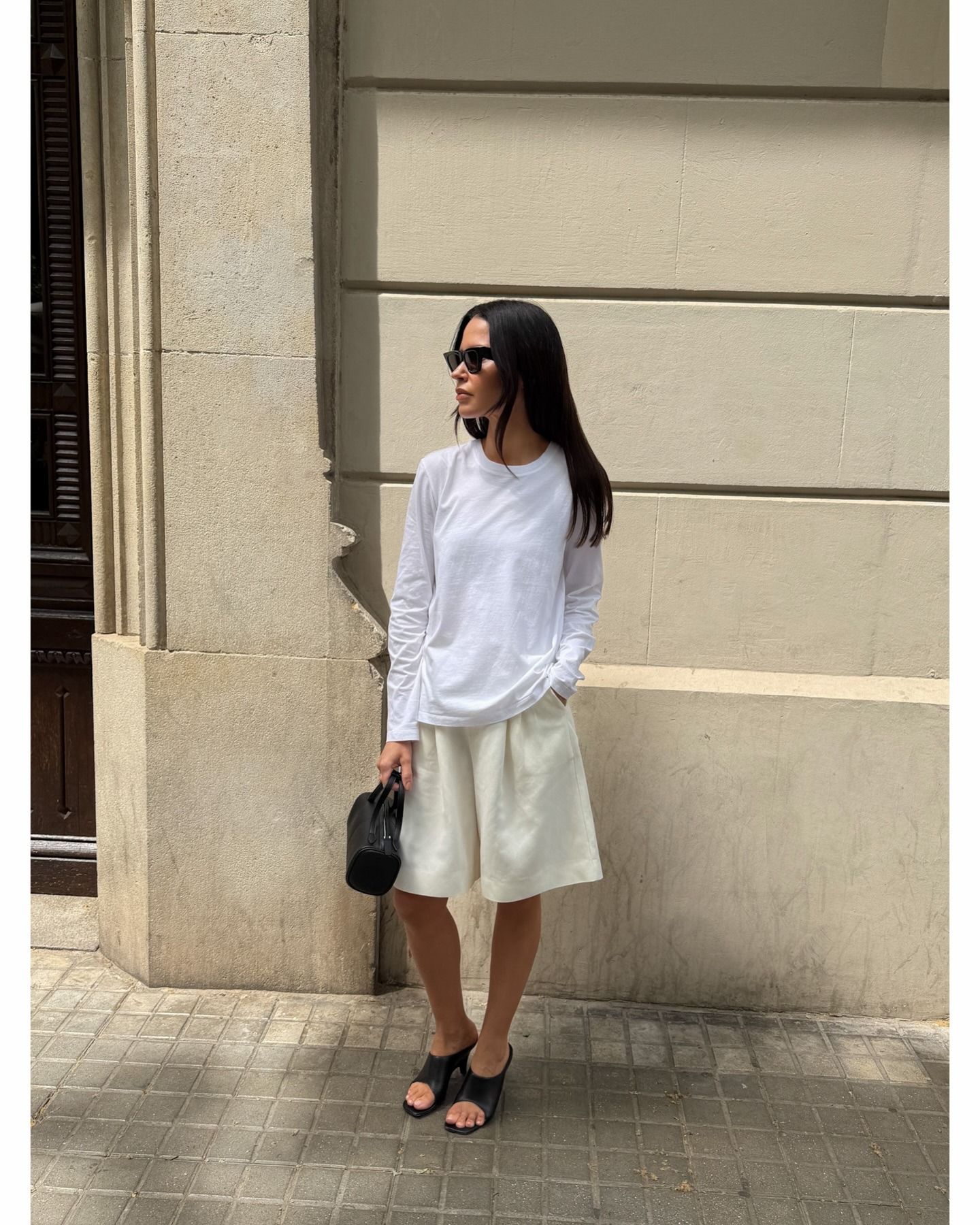 Johanna wears a white long-sleeve shirt, bermuda shorts, and black heels.