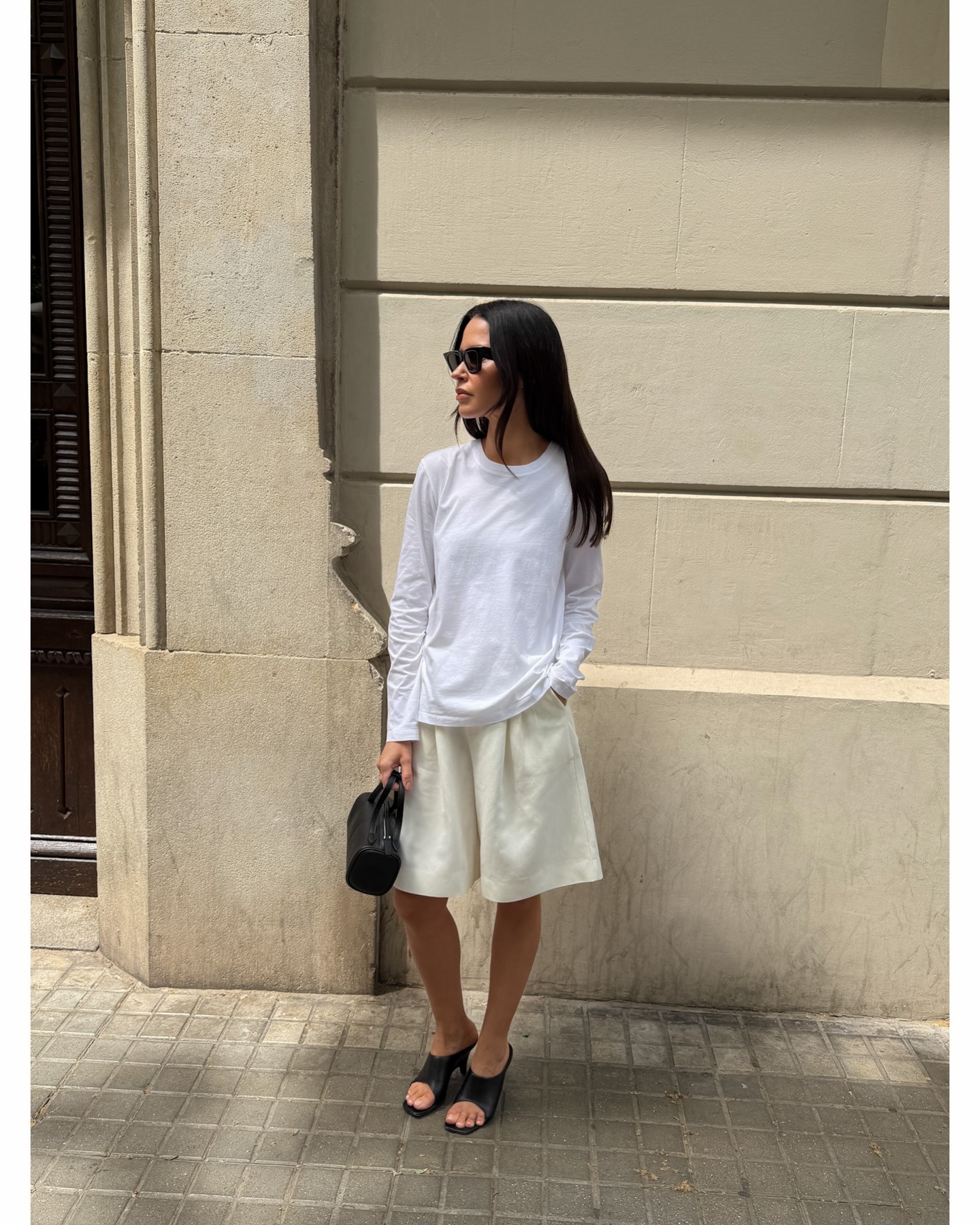 Johanna wears a white long-sleeved shirt, Bermuda shorts, and black heels.