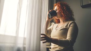 Woman looking out of the window, drinking a cup of coffee to deal with tiredness, one of the signs of high cortisol in women