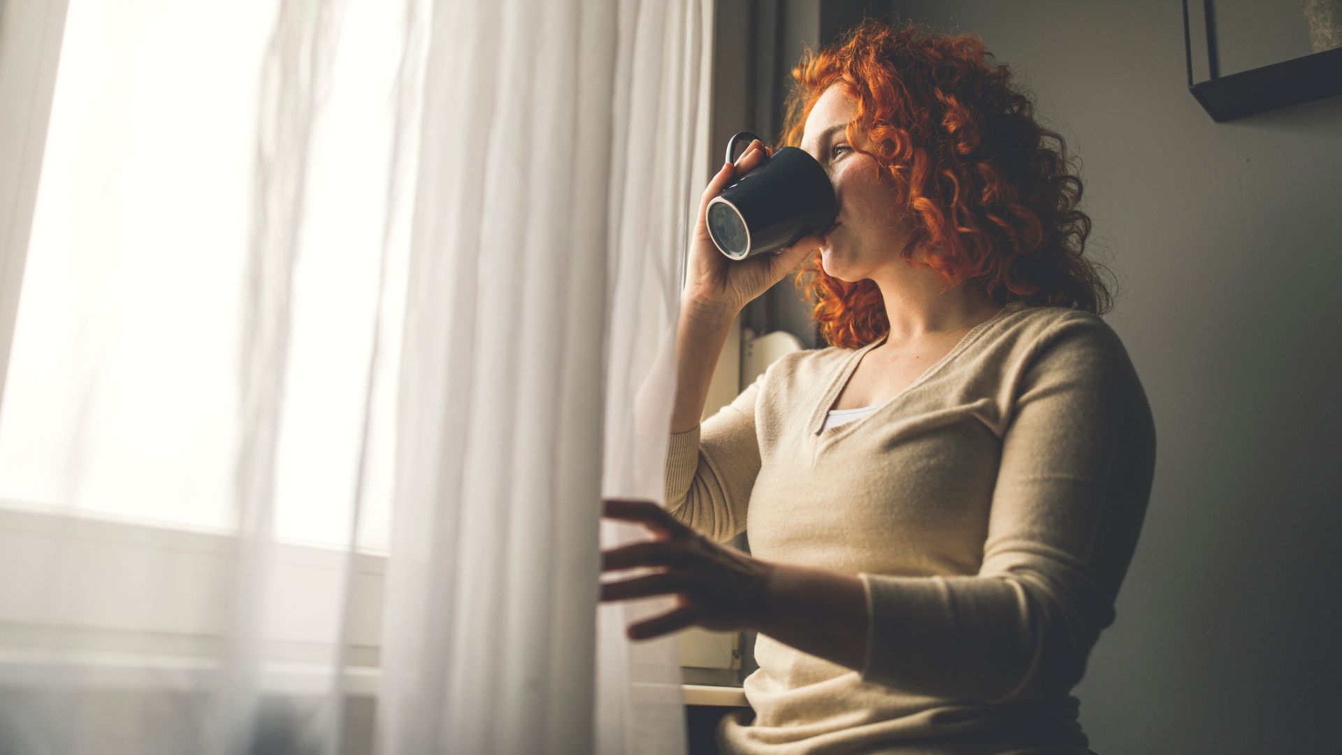 Žena, která se dívá z okna a pije šálek kávy, aby se vyrovnala s únavou, jedním z příznaků vysokého kortizolu u žen