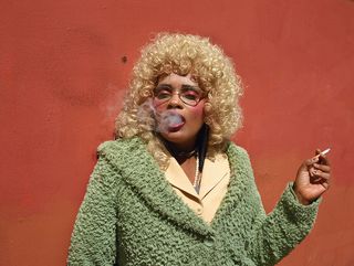 Woman with blond hair wearing green jacket smoking in front of orange wall
