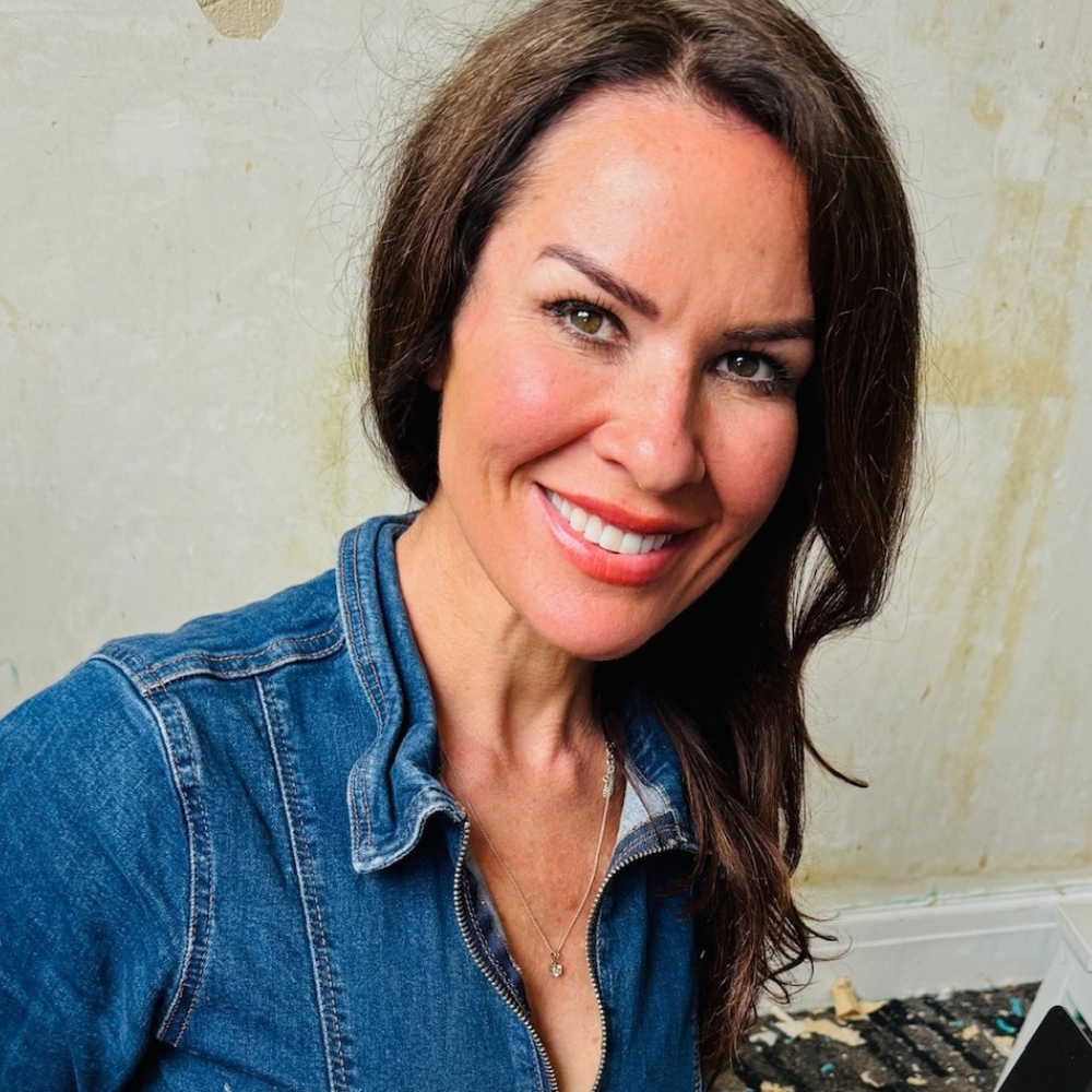 Smiling woman with long dark hair, wearing a denim jacket and necklace, against a weathered wall.