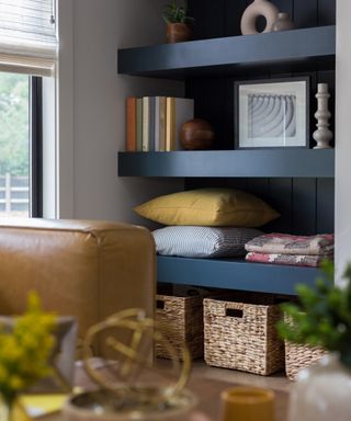 living room with detail of custom floating shelving in alcove next to fire place, baskets, couch pillows and books on shelves, corner of tan couch, vases on coffee table