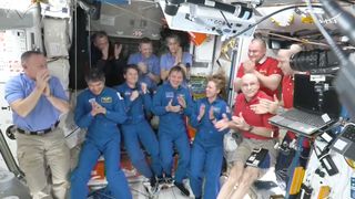 11 people — four in blue flight suits and the others in collared shirts and khaki pants — float on the international space station