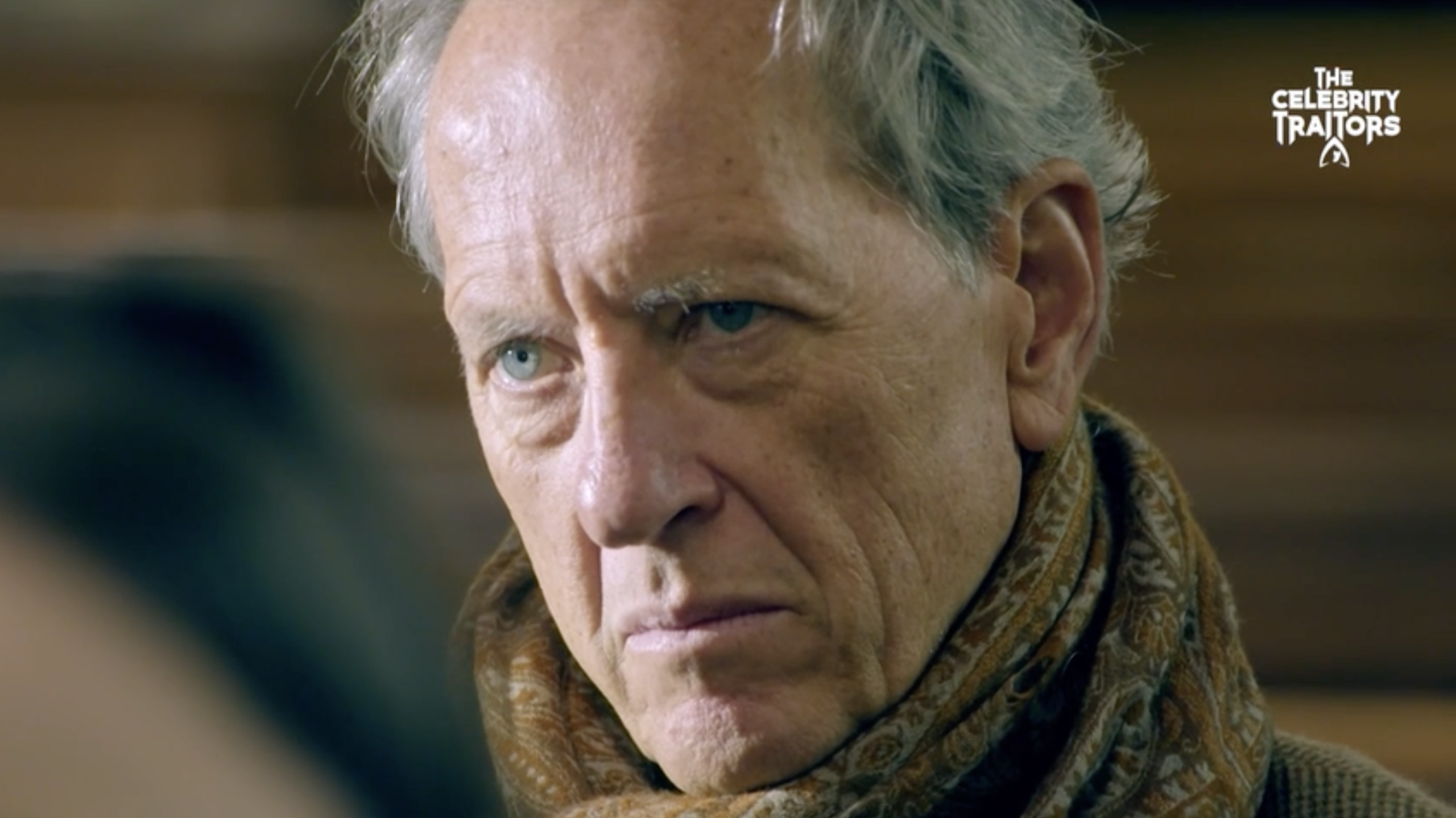 Close up of actor Richard E. Grant, wearing a brown patterned scarf, in the streaming trailer for the hit BBC TV show The Celebrity Traitors season 2 