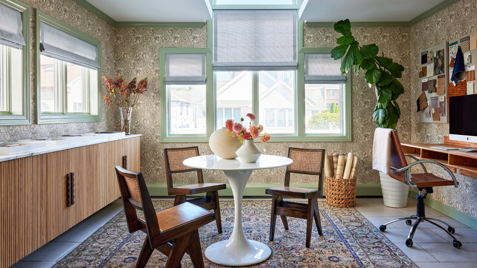 A light dining room with sage green trim, floral patterned wallpaper, a wooden bar, a white round dining table and three chairs, and a wooden desk