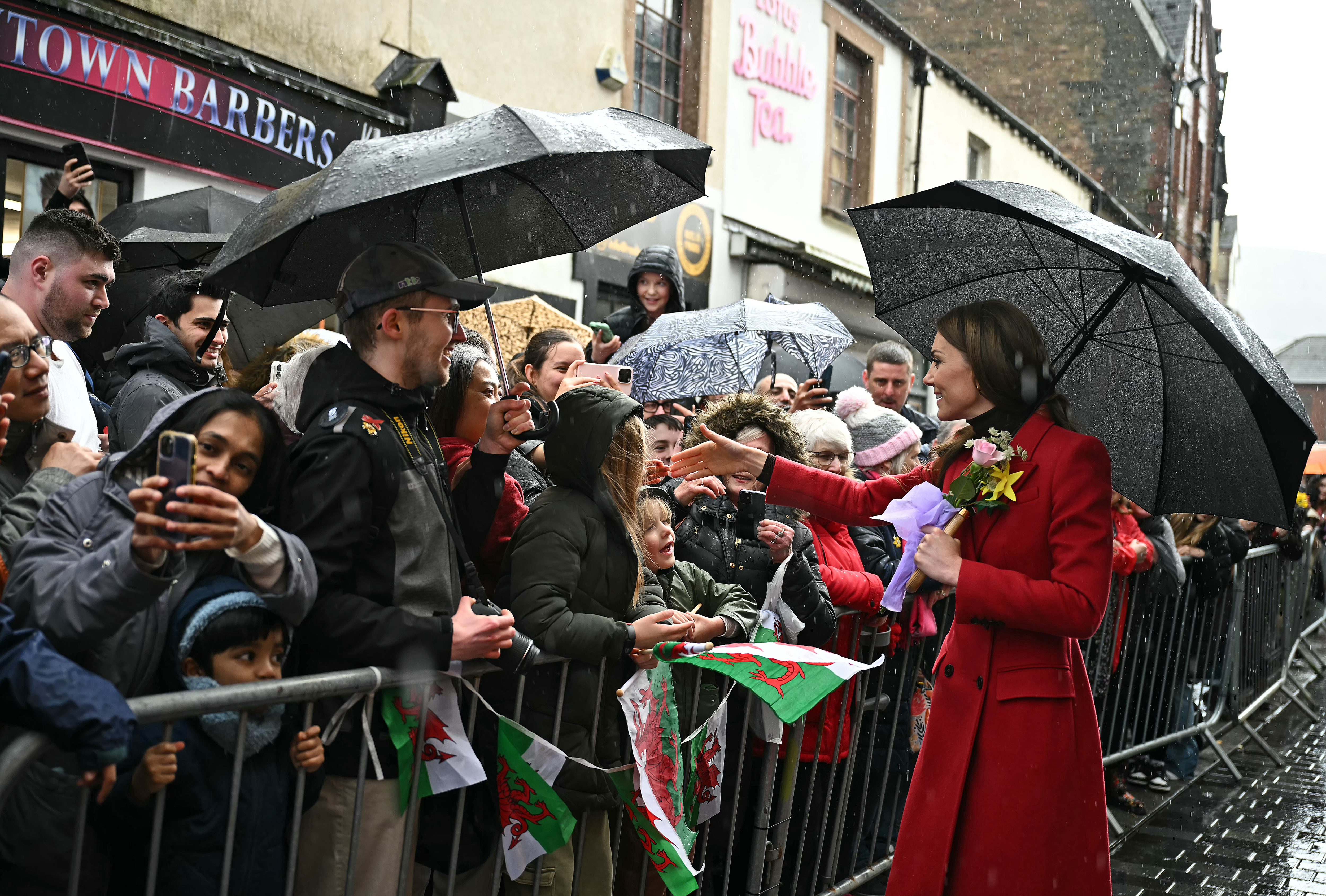Kate Middleton wearing a red coat and holding a black umbrella talking to a crowd of fans