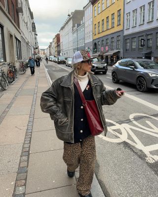 Sofia wears a brown leather jacket, a leopard hat, black cardigan, white turtleneck, red bag, leopard jeans, and black boots.