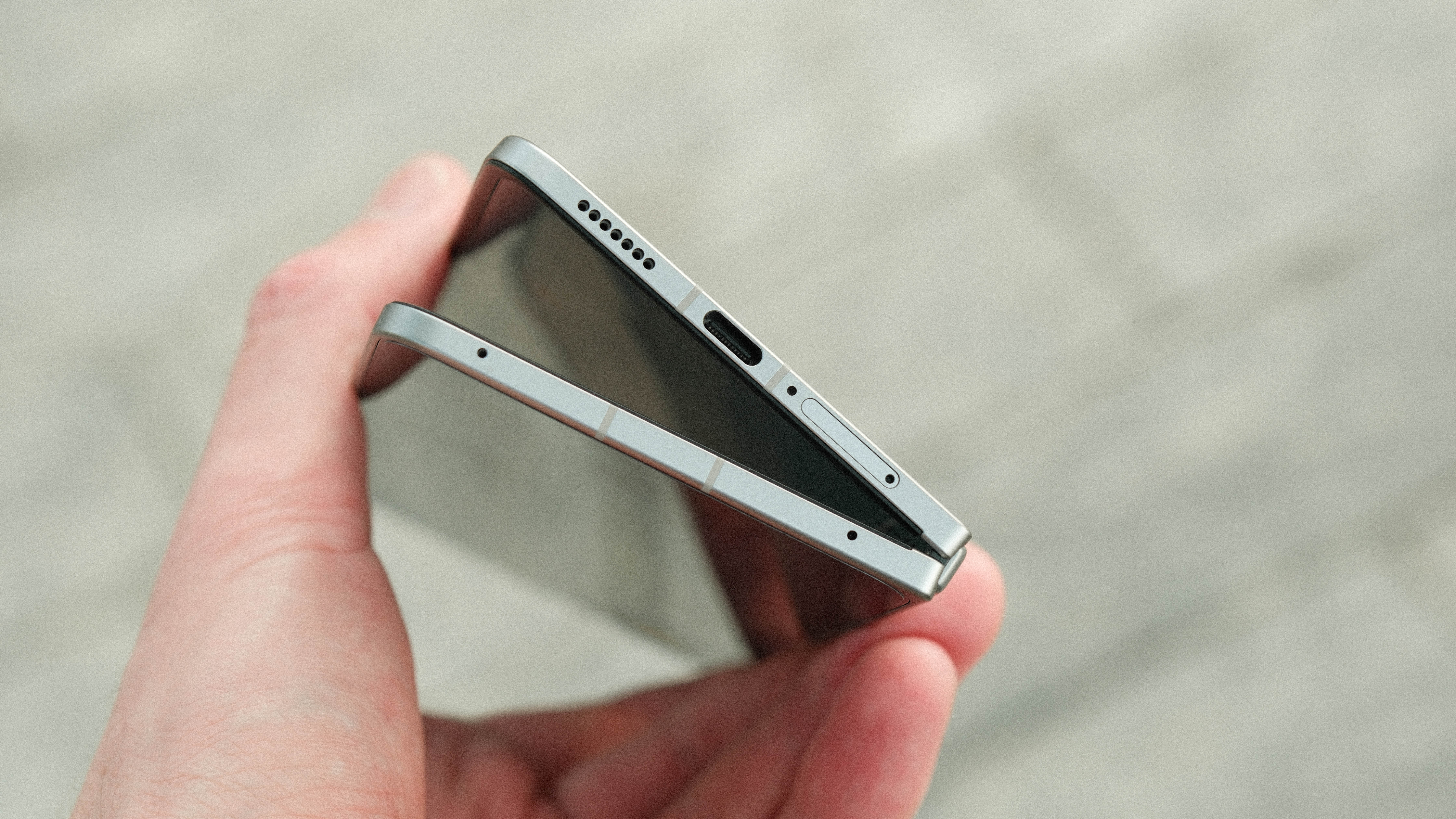 A close-up of the phone's bottom edge while partially folded in a V-shape. The USB-C port, speaker grille, and SIM tray are visible against a blurred stone background.