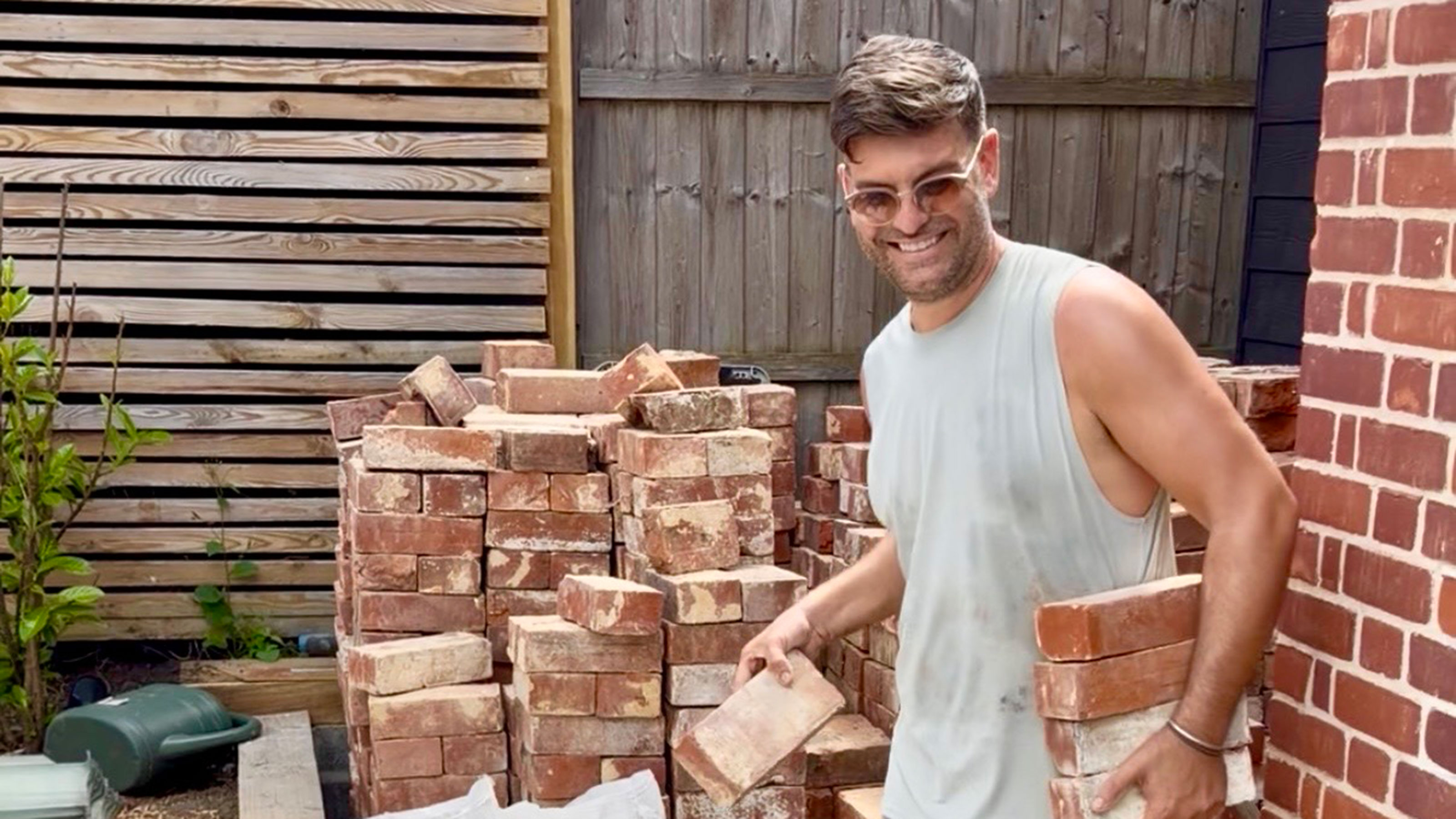 Man carrying bricks beside large pile of bricks by wooden fence in garden