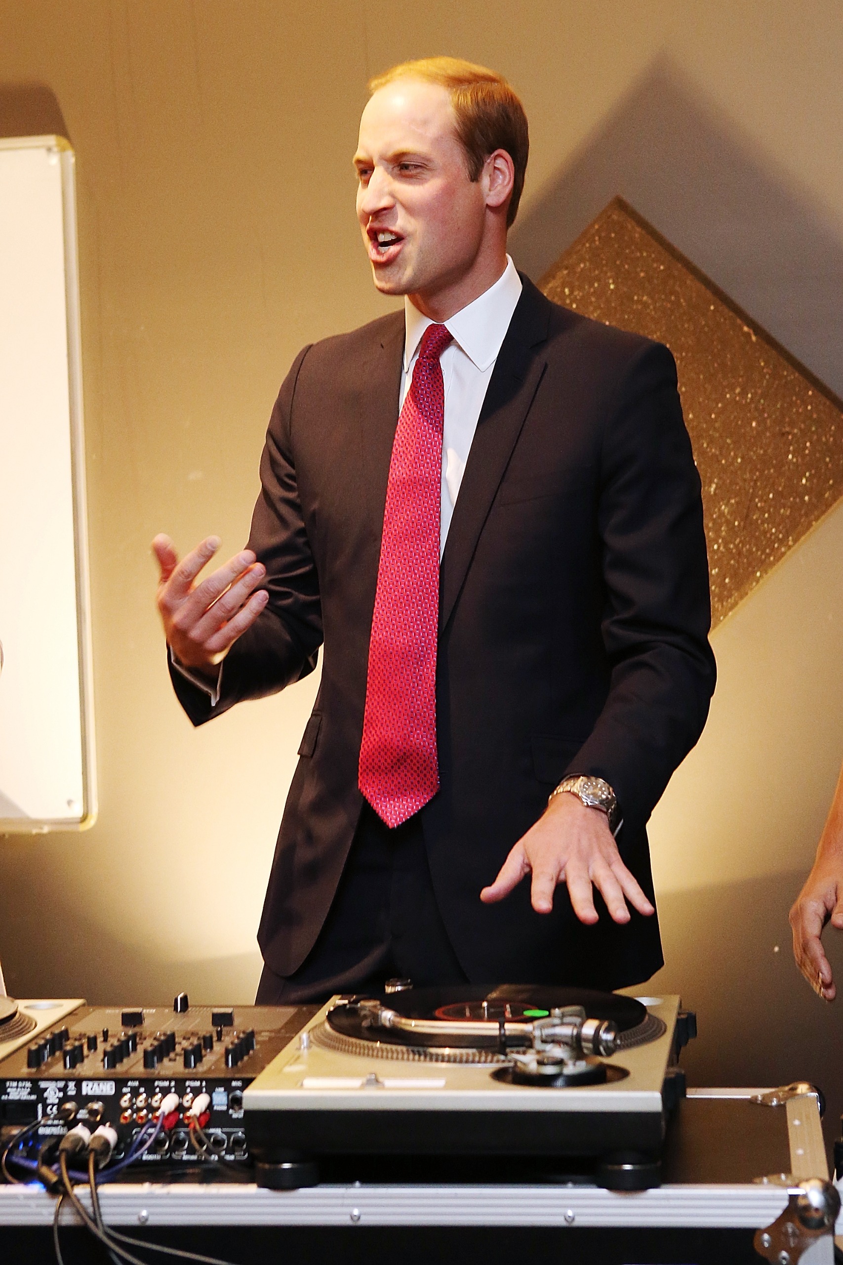 Prince William wearing a suit and tie standing at a DJ table