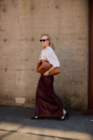 A woman wearing a white button-down short sleeve shirt, burgundy maxi skirt, black heels, and a oversized tan clutch bag.