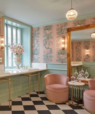 A bathroom with two white dual sinks and mint green paneling underneath, a gold floor mirror, two pink chairs with a side table in between, and light pink and green patterned wallpaper