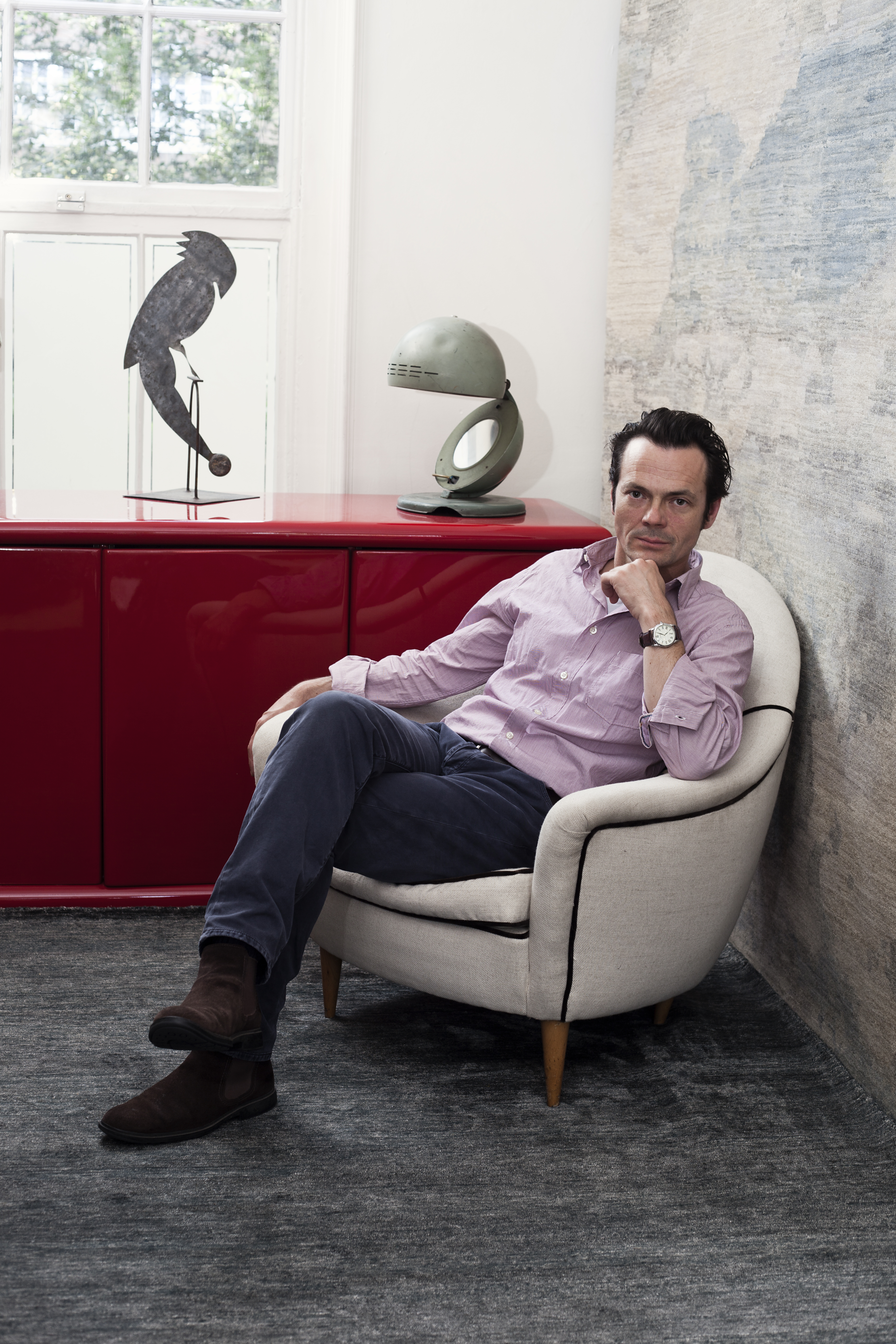 A man dressed in a long-sleeved pink shirt with blue trousers and brown shoes sits on a beige armchair in a rug-clad room with a lacquered red cabinet and two sculptural collectibles behind him.