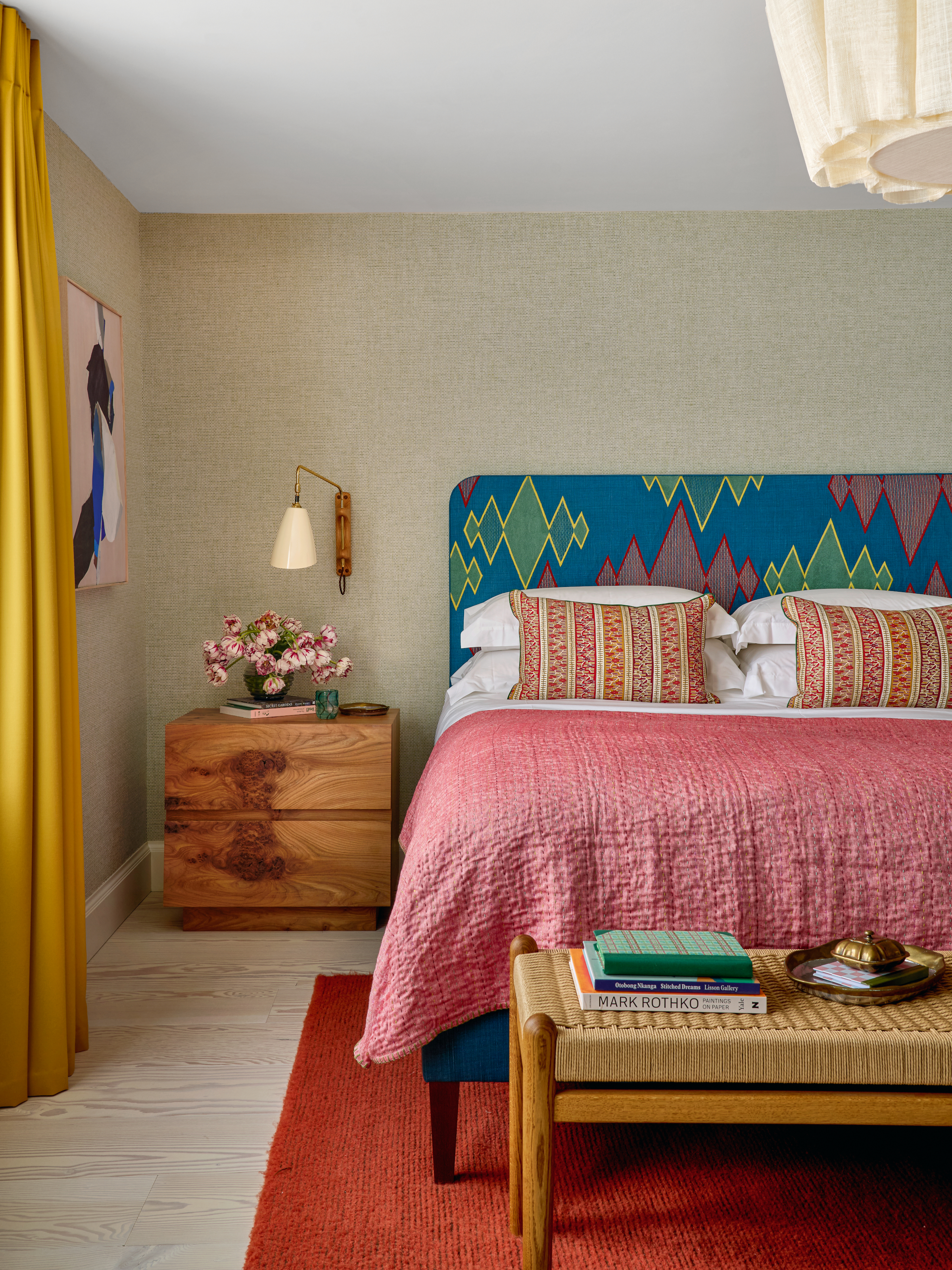 Bedroom with grasscloth wallpaper, blue geometric headboard, pink throw and wood bedside table
