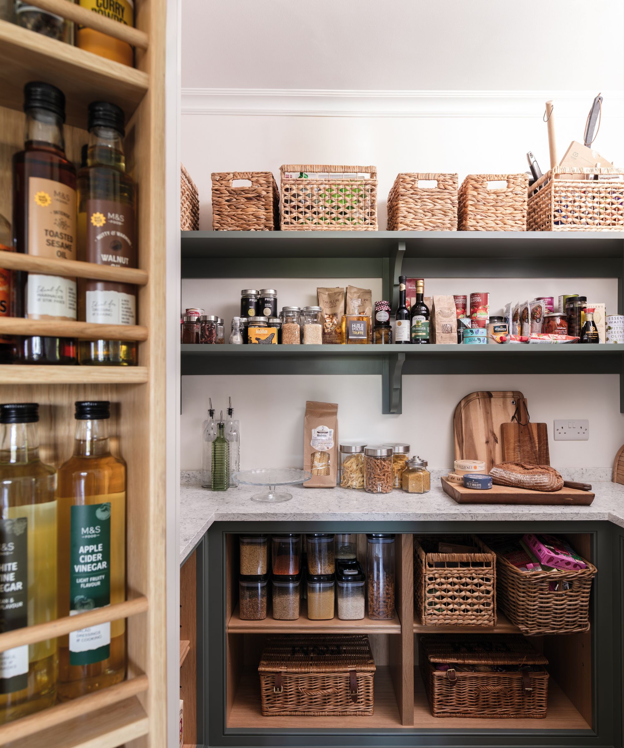 Well-organised walk-in pantry with dark shelving, woven storage baskets, labelled jars, and countertop prep space, blending practicality with relaxed, modern style.