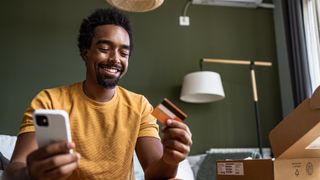 A man sits down on the sofa with a smile on his face, holding a credit card and a phone