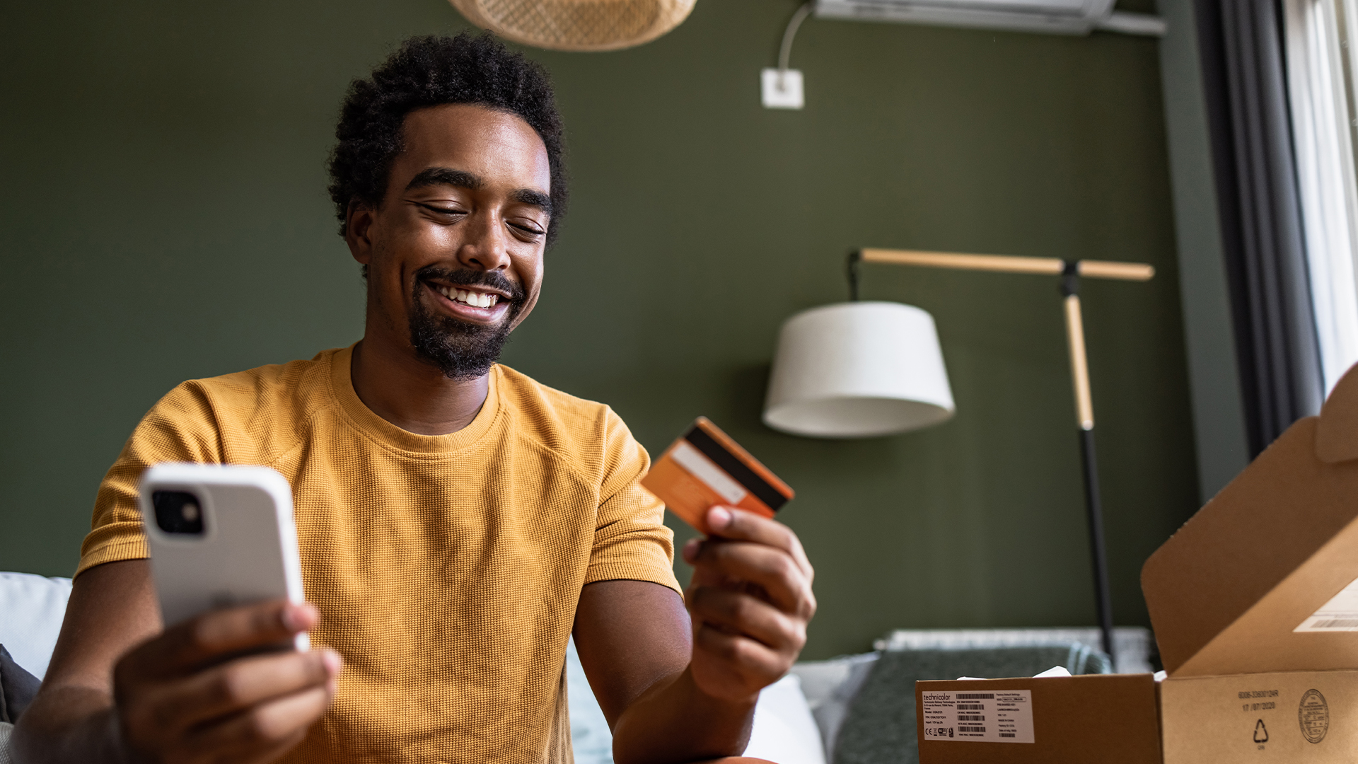 A man sits down on the sofa with a smile on his face, holding a credit card and a phone