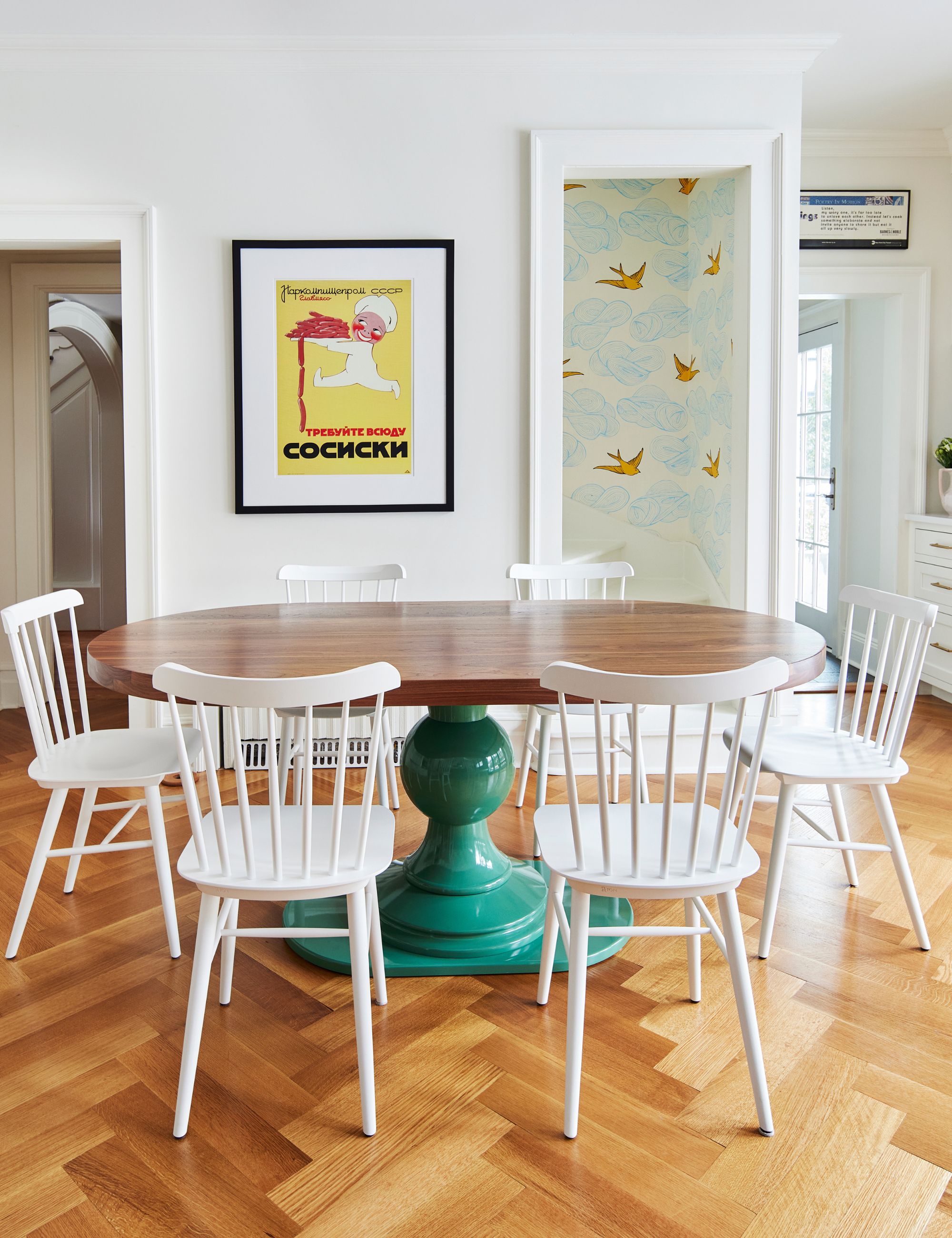 White dining room with wooden flooring, larger wooden dining table with green legs and white spindle-back chairs