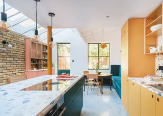 A colorful kitchen with orange cabinets on one side and green cabinets on the other side. There is also a brick wall on the far left of the kitchen.