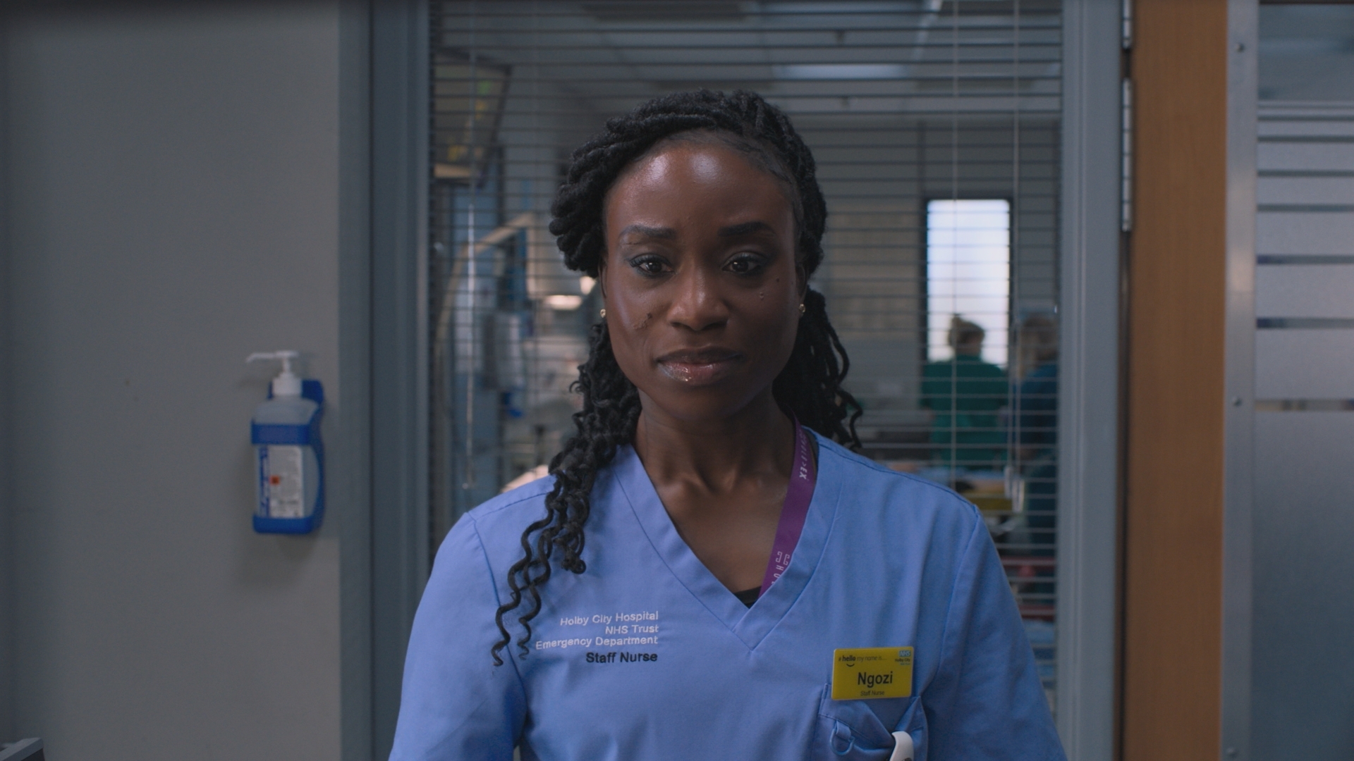 Ngozi at work looking straight forward with the hustle and bustle of Holby ED behind her in the background.