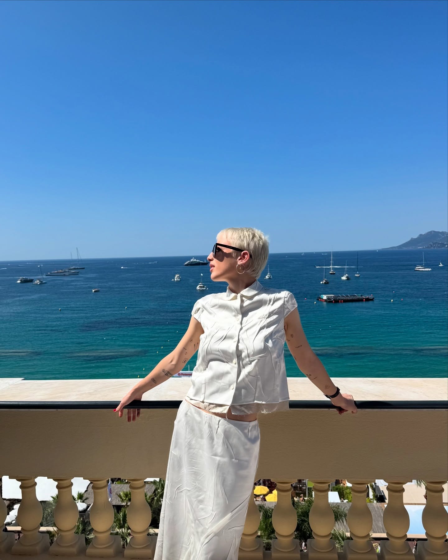 Emma Chamberlain leans on a railing in front of the ocean wearing a white short sleeve blouse and a white cotton skirt.