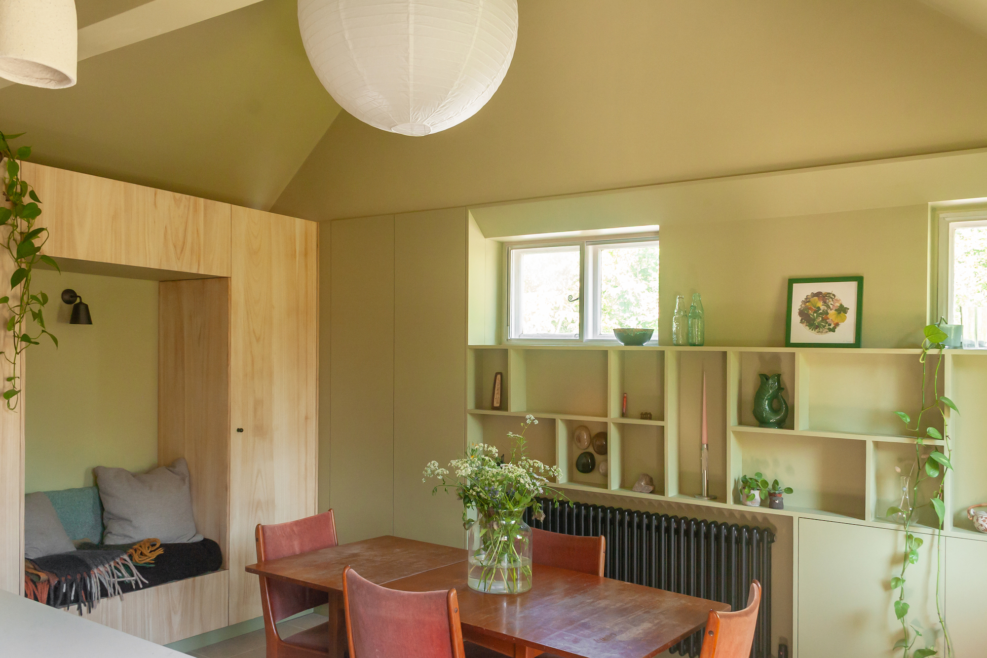 A view of the dining area with the built in bench seat in the nook on the far left wall, the walls are colour drenched in a sage/avocado green, including the wall shelves on the far right wall. There is a large light above the orange wood dining table.