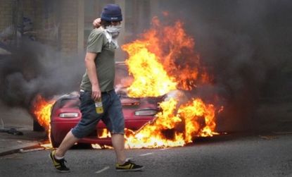 A masked man walks past a burning car in London