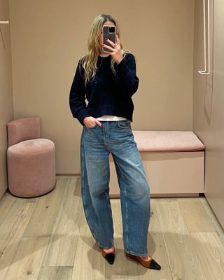 Fashion editor Eliza Huber poses for a changing room mirror selfie wearing a dark crewneck sweater layered over a white tee, barrel jeans, and pointed cap-toe shoes