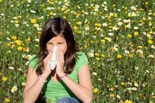 A woman sits near a field of flowers, blowing her nose.