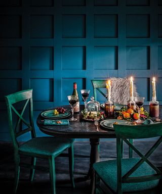 A dining room with a navy blue grid accent wall and a forest green dining room set with a table with tableware, wine glasses, cutlery, and candles, and three chairs around the table