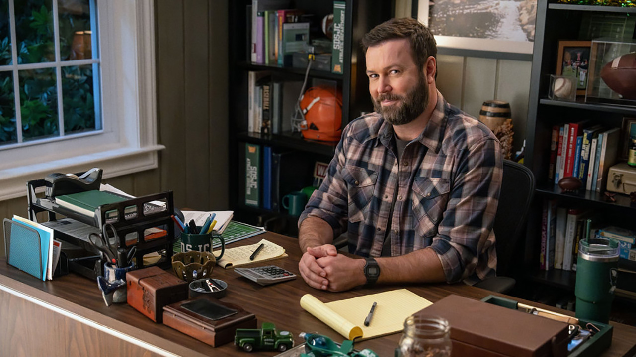 Taran Killam sits at his football desk attempting to look busy in a plaid shirt on Stumble.