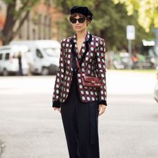 a woman at milan fashion week wearing a patterned silk blazer, black trousers, sunglasses and a beret