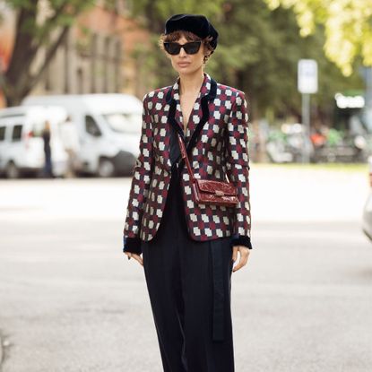 a woman at milan fashion week wearing a patterned silk blazer, black trousers, sunglasses and a beret