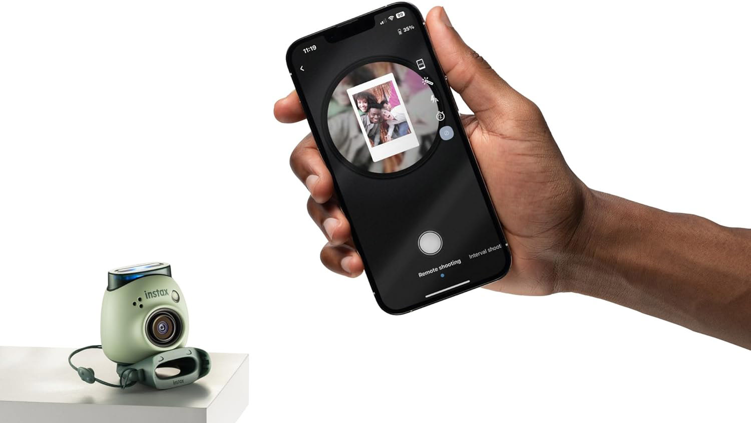 Fujifilm Instax Pal in green color on a white background. The Instax Pal is placed on a table on the left of the frame and there is a hand holding a smartphone on the right of the image, showing the Fujifilm Instax Pal app in use